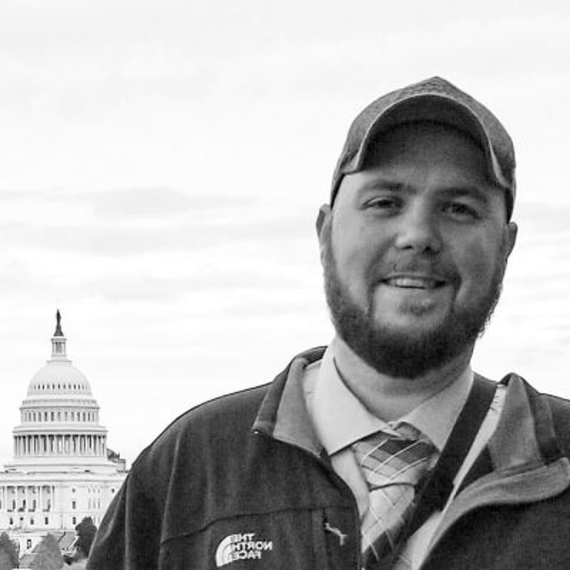 The image shows a man in front of the United States Capitol Building. He is wearing a cap, a jacket, and a tie. He has a beard and is smiling. The background is a cloudy sky.